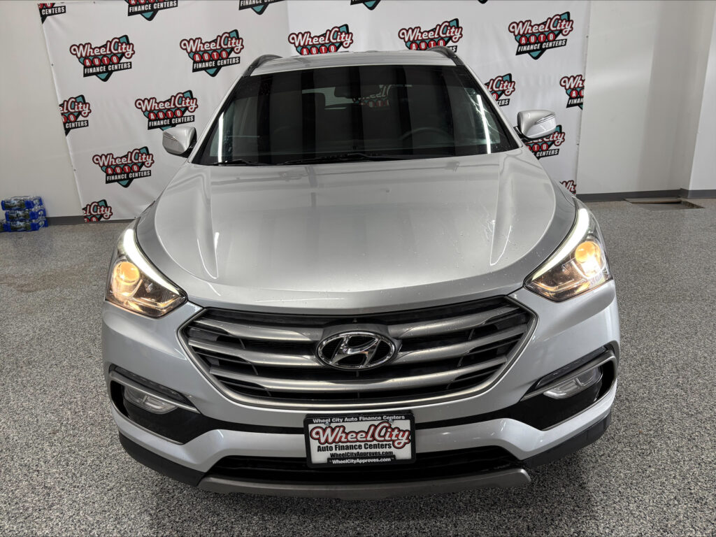 Front view of a silver Hyundai SUV in a showroom with a backdrop covered in Wheel City Auto Finance Centers logos; headlights on.