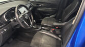 Interior of a blue car showing the driver's seat, steering wheel, center console, and front passenger seat with black upholstery from the open door side view.