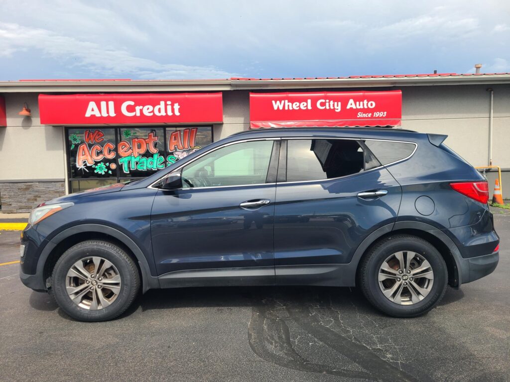 Blue SUV parked in front of a storefront with red All Credit and Wheel City Auto signs on the awnings. The window displays graphics reading 'We Accept All Trade'.