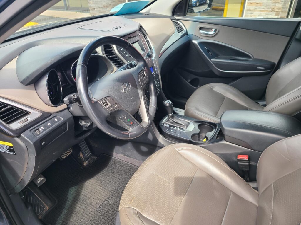 Interior view of a car's driver side: steering wheel with control buttons, dashboard, and center console including gear shifter and cup holders.