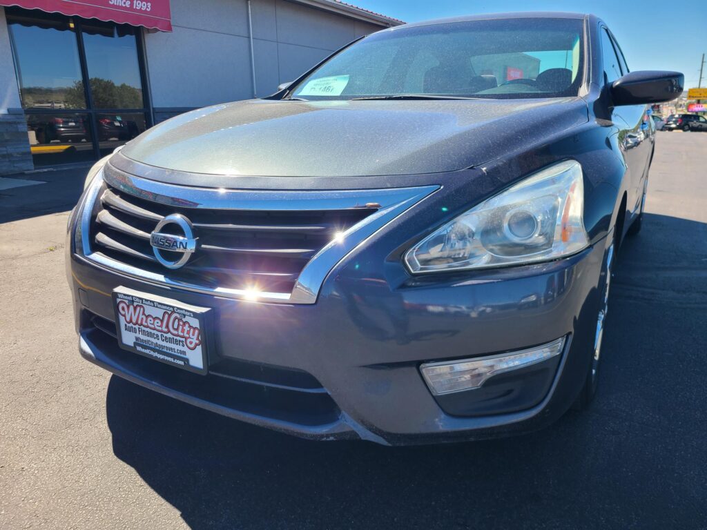 Front three-quarter view of a dark gray Nissan SUV parked at a dealership, showing a Wheel City Auto Finance license plate frame on the front bumper and a vehicle info sticker on the windshield.