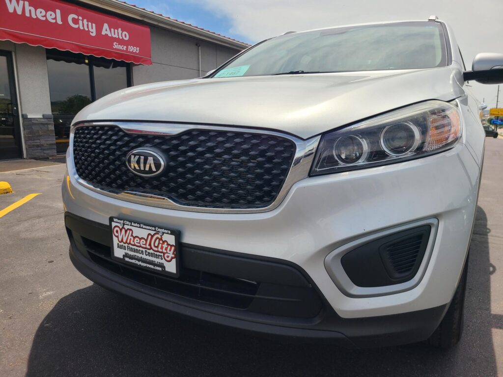 Front view of a silver Kia SUV parked at a Wheel City Auto dealership, showing the grille, headlights, and Wheel City license plate frame