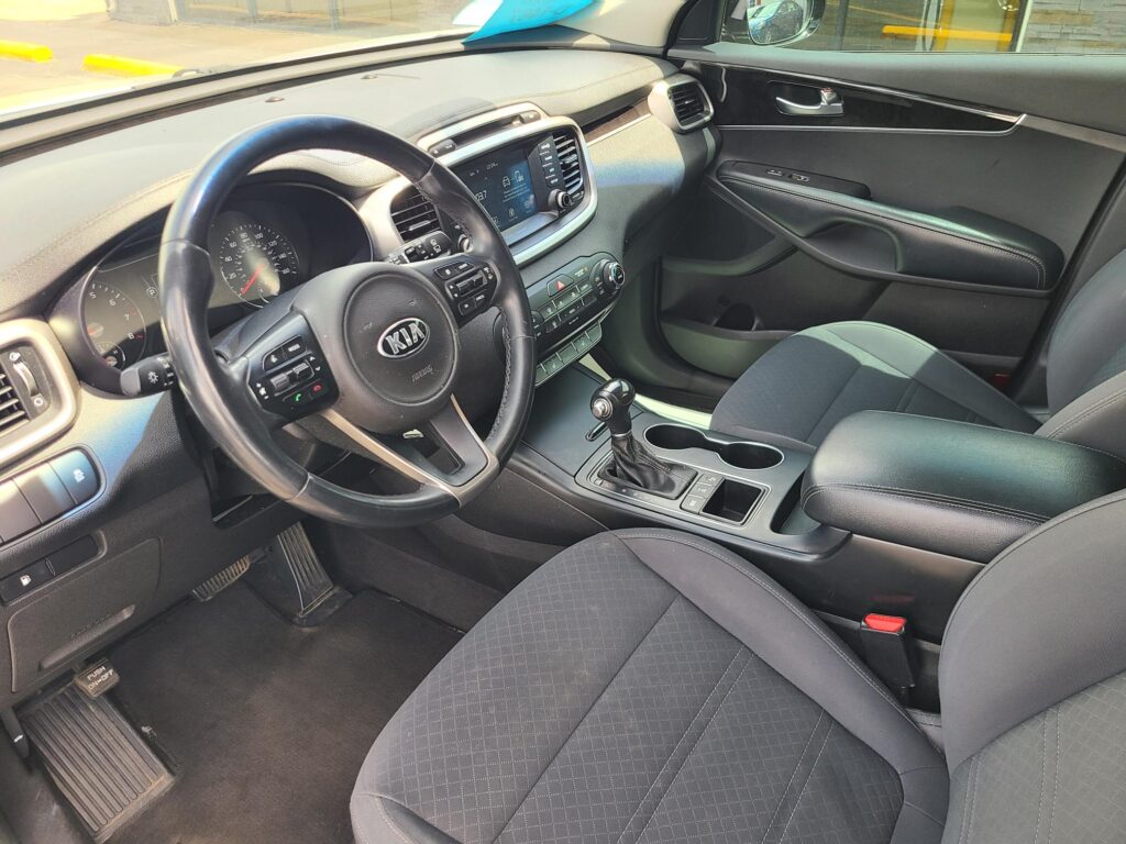 Front interior of a Kia SUV showing steering wheel, center touchscreen, gear shifter, and black cloth seats in a clean cabin.