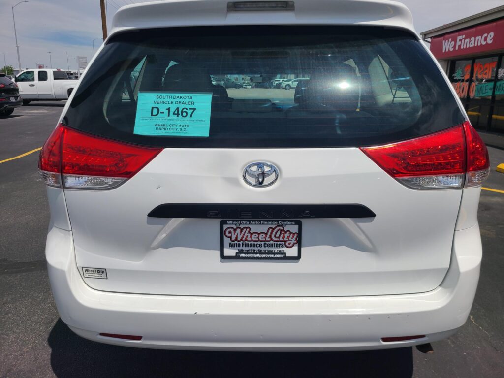 Rear view of a white Toyota Sienna with a teal dealer placard visible in the rear window showing South Dakota Vehicle Dealer D-1467 and Wheel City Auto Finance Centers groundplate area.