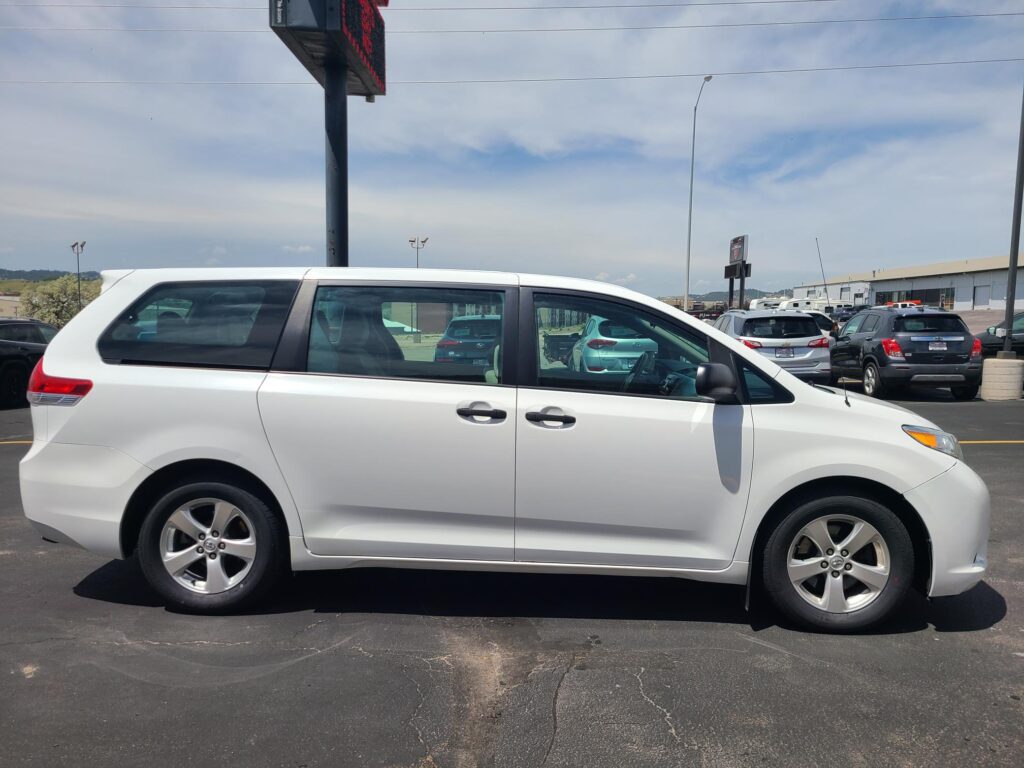 Side view of a white minivan parked in a car dealership lot with other vehicles and a building in the background.