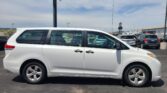 Side view of a white minivan parked in a car dealership lot with other vehicles and a building in the background.