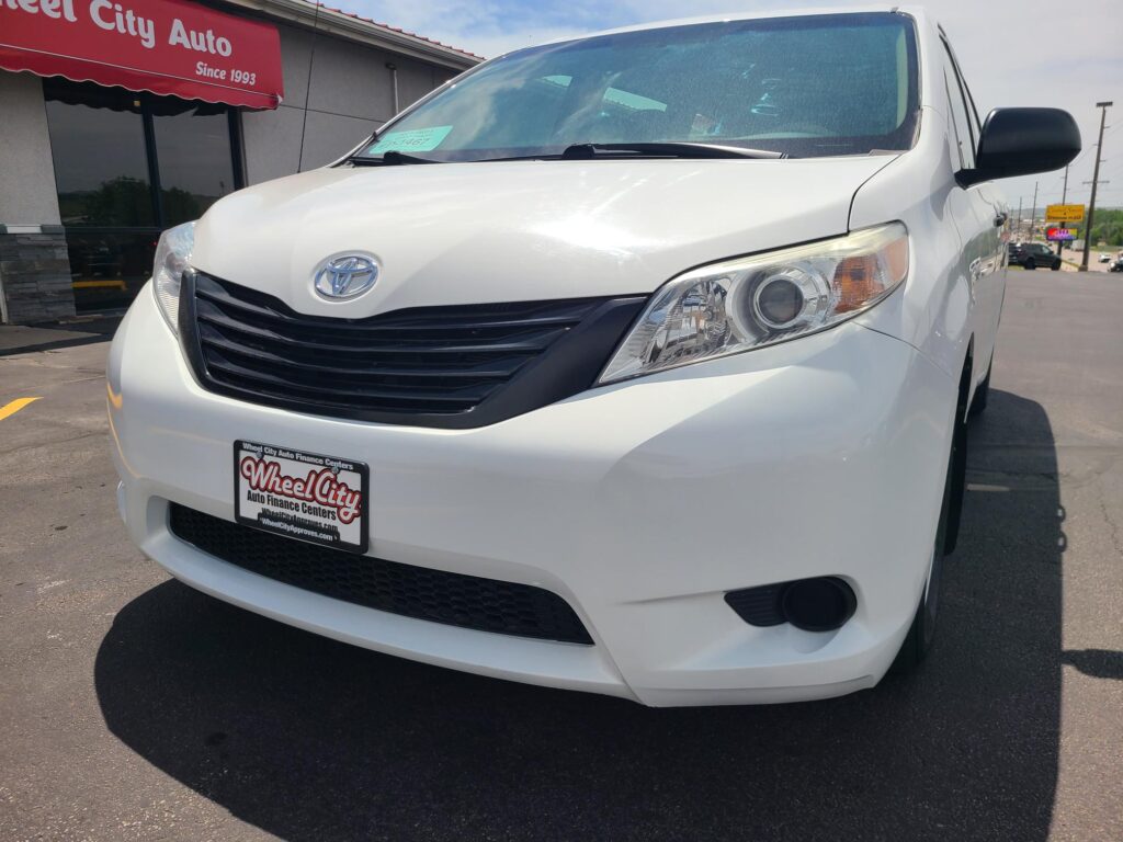 White Toyota van at Wheel City Auto dealership, front grille and headlight visible with a red dealership awning in the background.