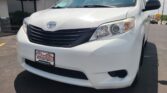 White Toyota van at Wheel City Auto dealership, front grille and headlight visible with a red dealership awning in the background.