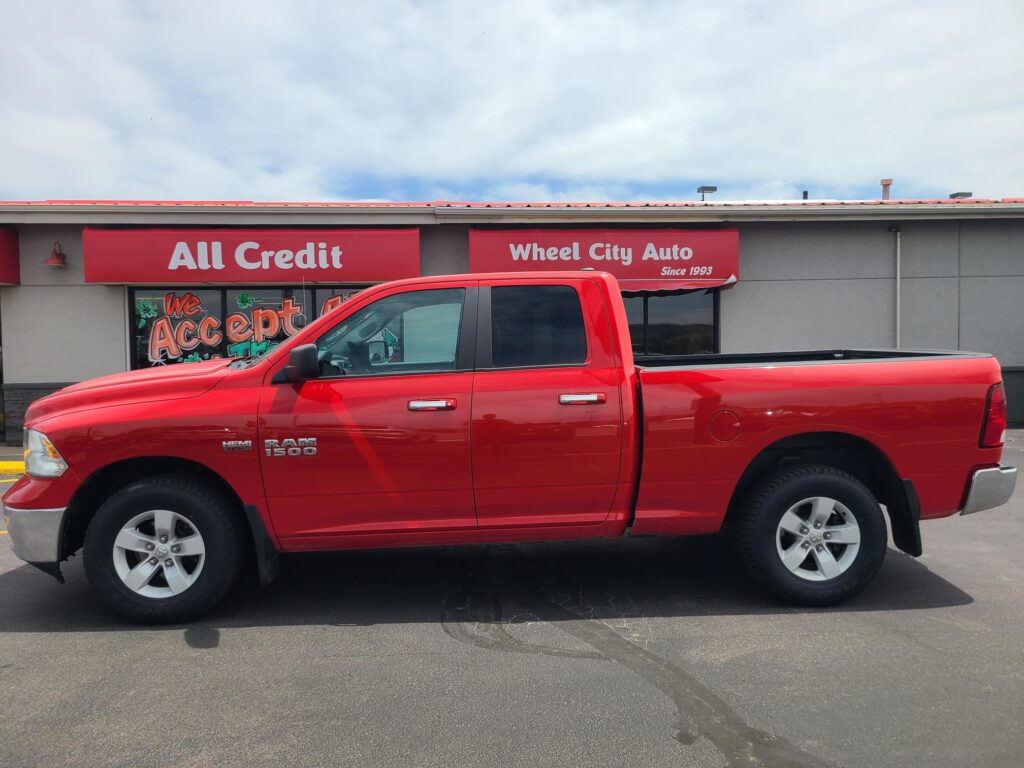 Red RAM 1500 pickup parked in a parking lot in front of a gray storefront with red All Credit and Wheel City Auto signs.