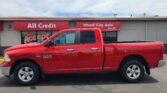 Red RAM 1500 pickup parked in a parking lot in front of a gray storefront with red All Credit and Wheel City Auto signs.