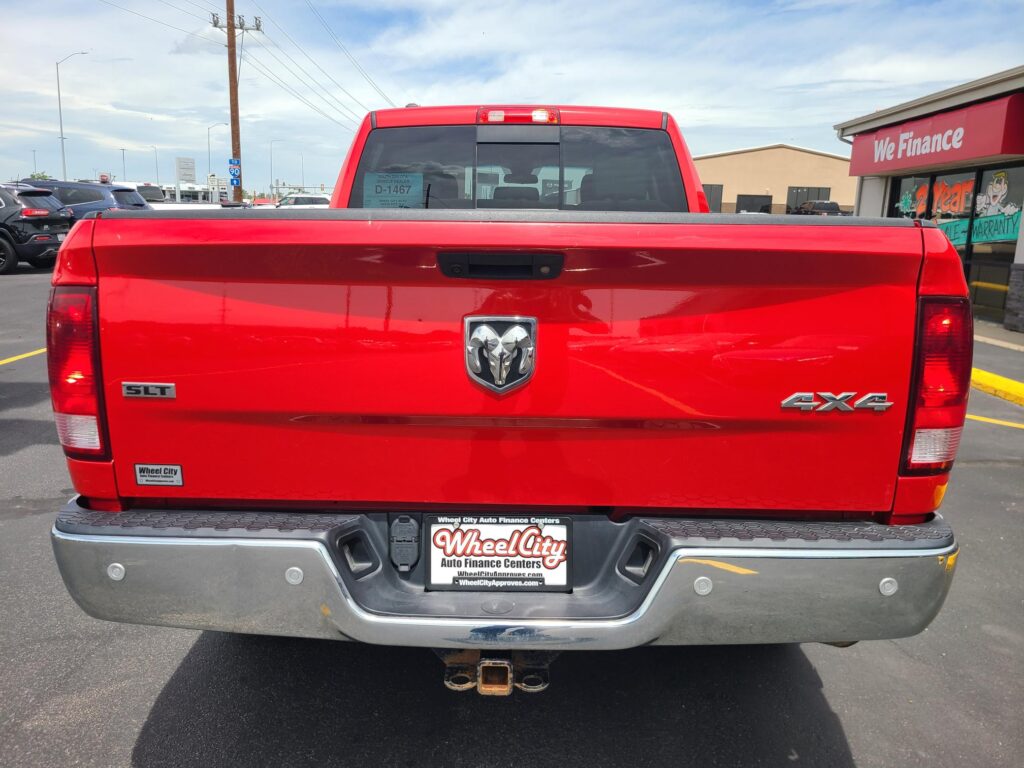 Rear view of a red pickup truck in a parking lot, showing the Ram emblem on the tailgate, a tow hitch, and Wheel City dealership plate frame near the bumper.