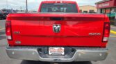 Rear view of a red pickup truck in a parking lot, showing the Ram emblem on the tailgate, a tow hitch, and Wheel City dealership plate frame near the bumper.