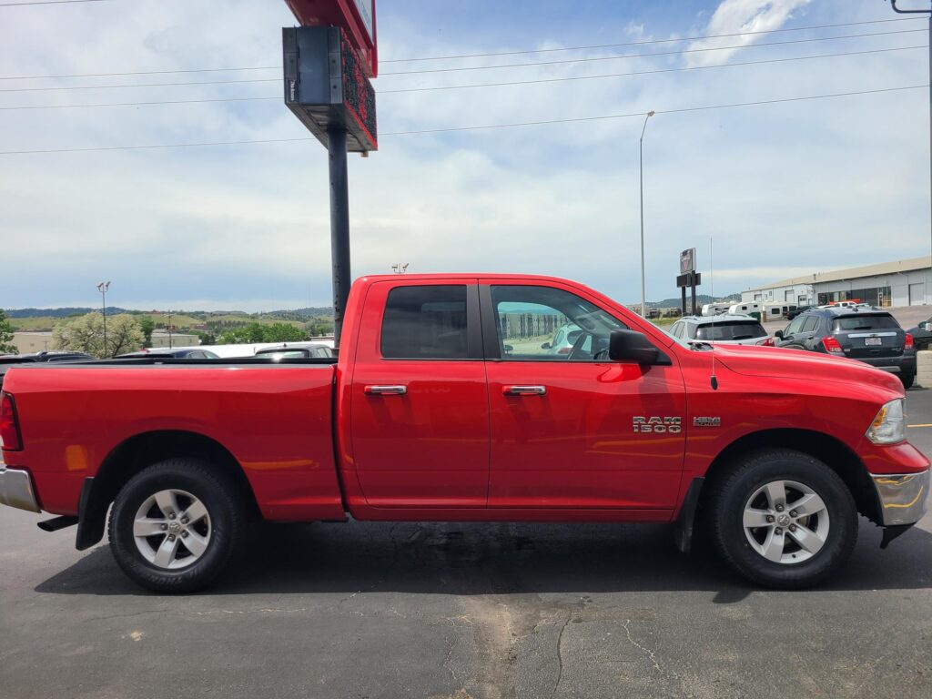 Bright red Ram 1500 pickup truck parked in a parking lot with other cars and a dealership in the background.