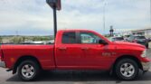 Bright red Ram 1500 pickup truck parked in a parking lot with other cars and a dealership in the background.