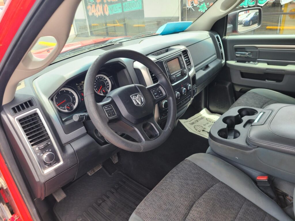 Interior of a red pickup truck showing the driver’s area: steering wheel, dashboard with infotainment screen, and center controls visible from the passenger seat angle.