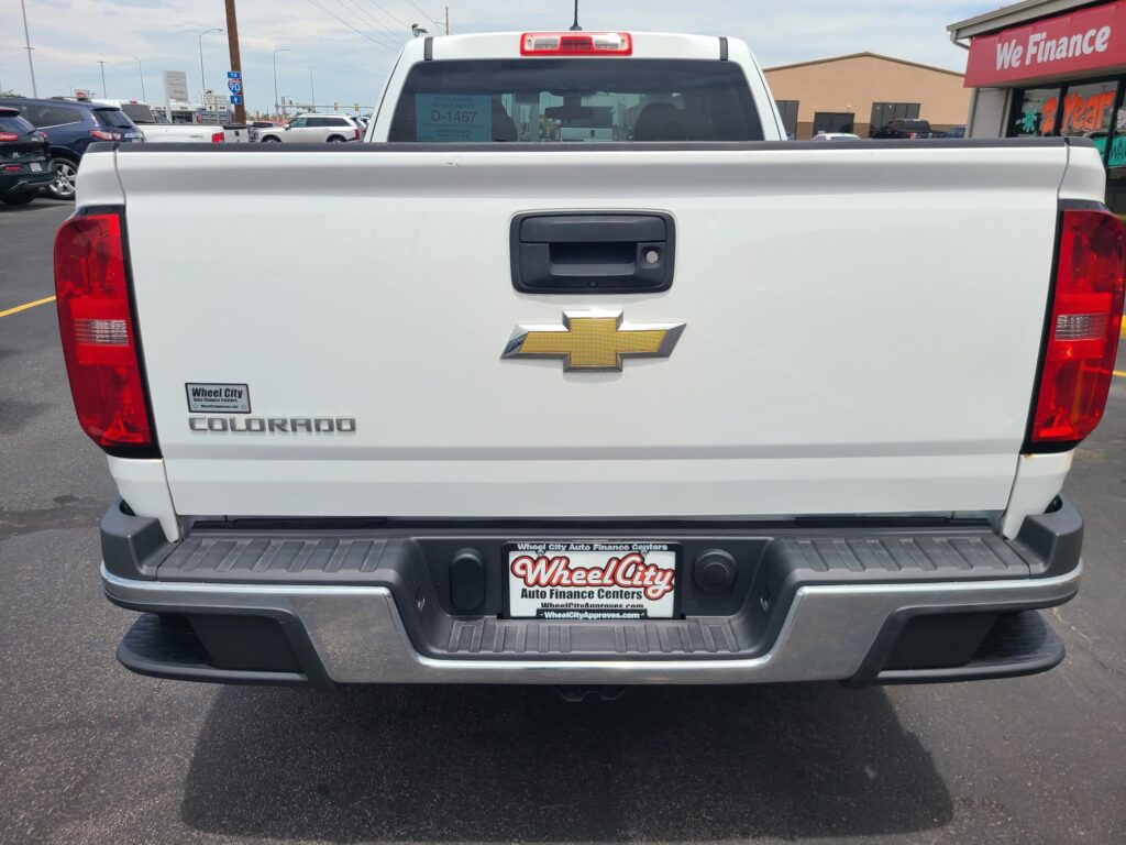 Rear view of a white Chevrolet Colorado pickup in a car dealership lot, with a Wheel City license plate frame and red tail lights visible.