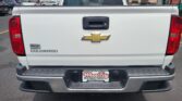 Rear view of a white Chevrolet Colorado pickup in a car dealership lot, with a Wheel City license plate frame and red tail lights visible.