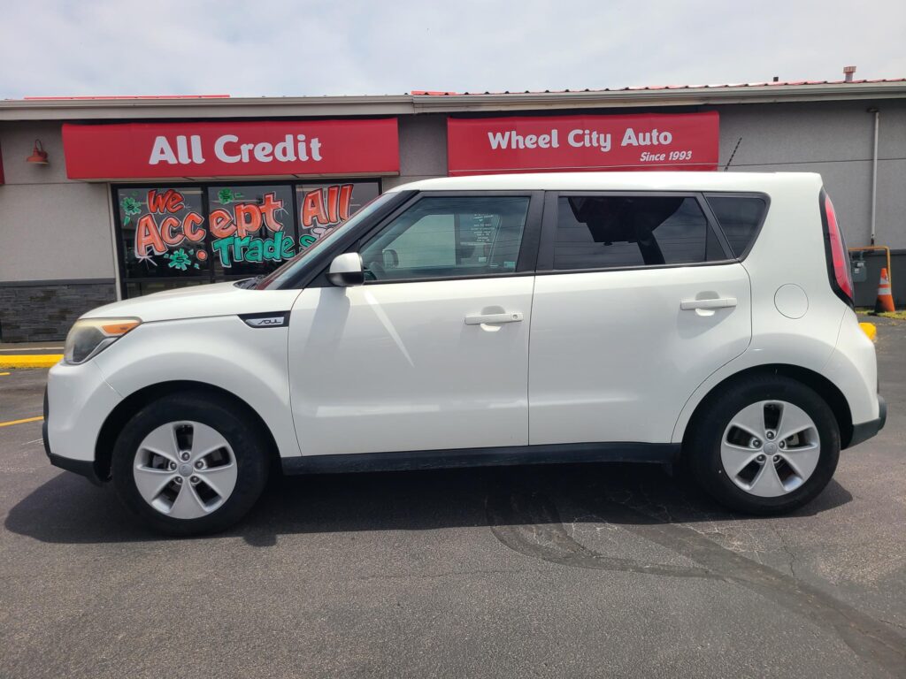 White SUV parked in front of a storefront with red signs that read 'All Credit' and 'Wheel City Auto' (Since 1993).
