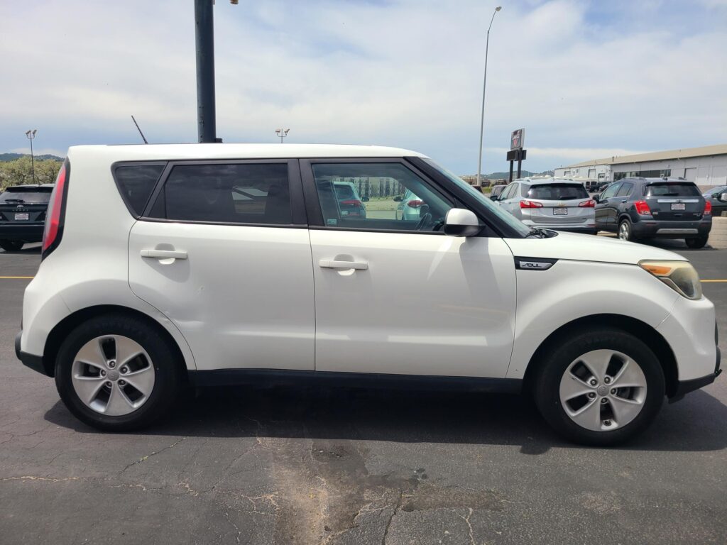 Side view of a white Kia Soul parked in a lot with other cars and a commercial building in the background.