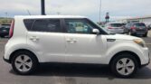 Side view of a white Kia Soul parked in a lot with other cars and a commercial building in the background.