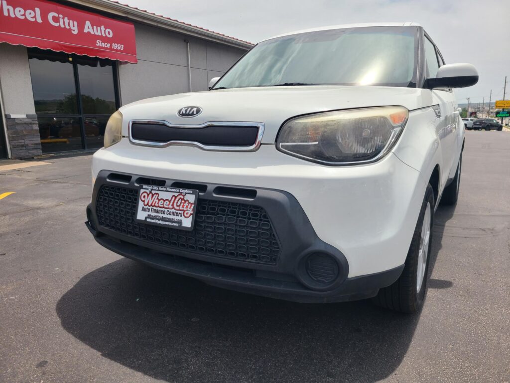White Kia van parked in front of a dealership with a red canopy reading 'Wheel City Auto Since 1993'.