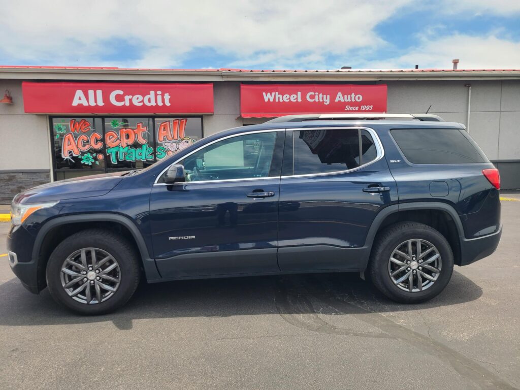 Blue GMC Acadia SUV parked in front of a storefront with red banners reading All Credit and Wheel City Auto; window sign reads We Accept All Trades.