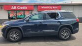 Blue GMC Acadia SUV parked in front of a storefront with red banners reading All Credit and Wheel City Auto; window sign reads We Accept All Trades.