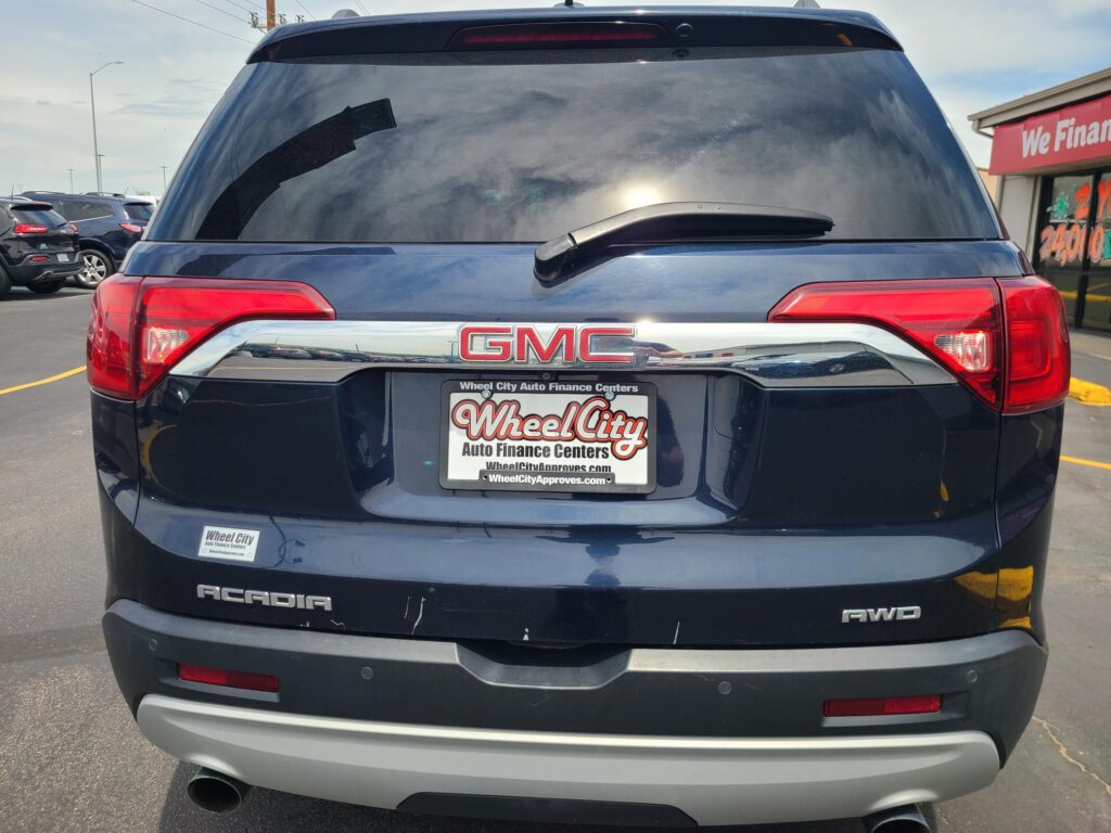 Rear view of a blue GMC Acadia SUV, showing the tailgate, rear windshield wiper, and red taillights in a car lot. License plate frame reads Wheel City Auto Finance Centers.