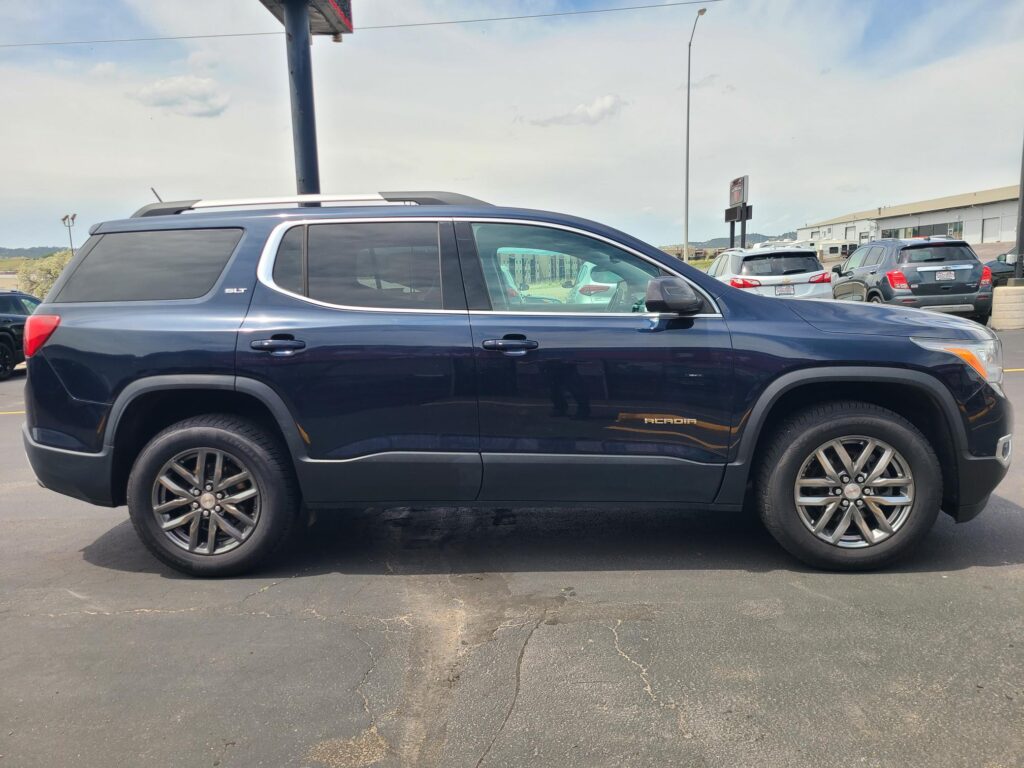 Side view of a dark blue GMC Acadia SUV parked in a lot, with other cars and a warehouse in the background, under a cloudy sky.