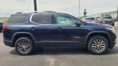Side view of a dark blue GMC Acadia SUV parked in a lot, with other cars and a warehouse in the background, under a cloudy sky.
