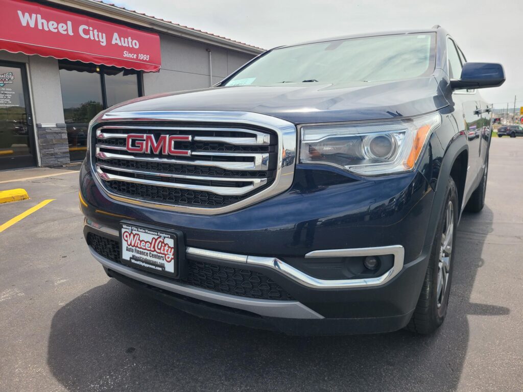 Front view of a dark gray GMC pickup truck parked in front of a red Wheel City Auto storefront awning with chrome grille and large GMC emblem.