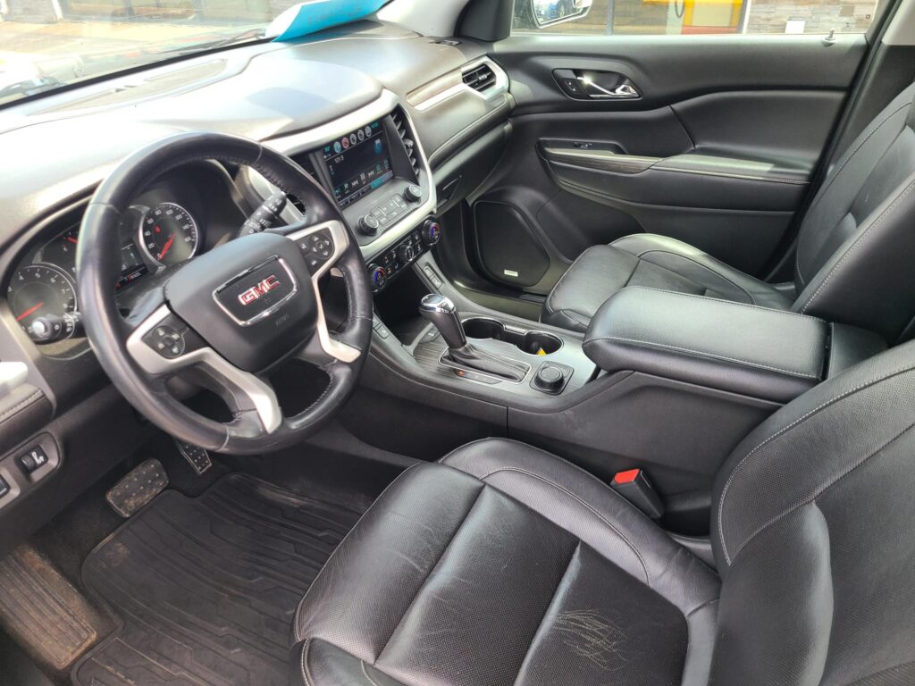 Interior of a GMC vehicle: driver’s seat, steering wheel, center touchscreen display, climate controls, and black leather upholstery with stitching.
