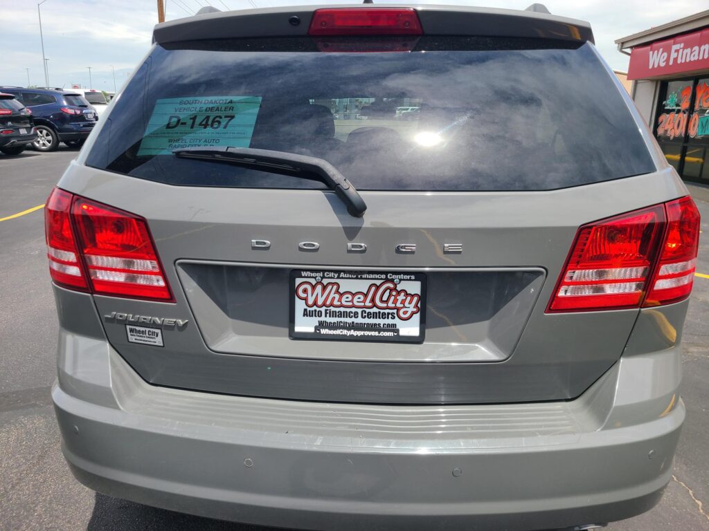 Rear view of a gray Dodge Journey SUV in a dealership lot, teal D-1467 decal visible on the rear window, Wheel City license plate frame.