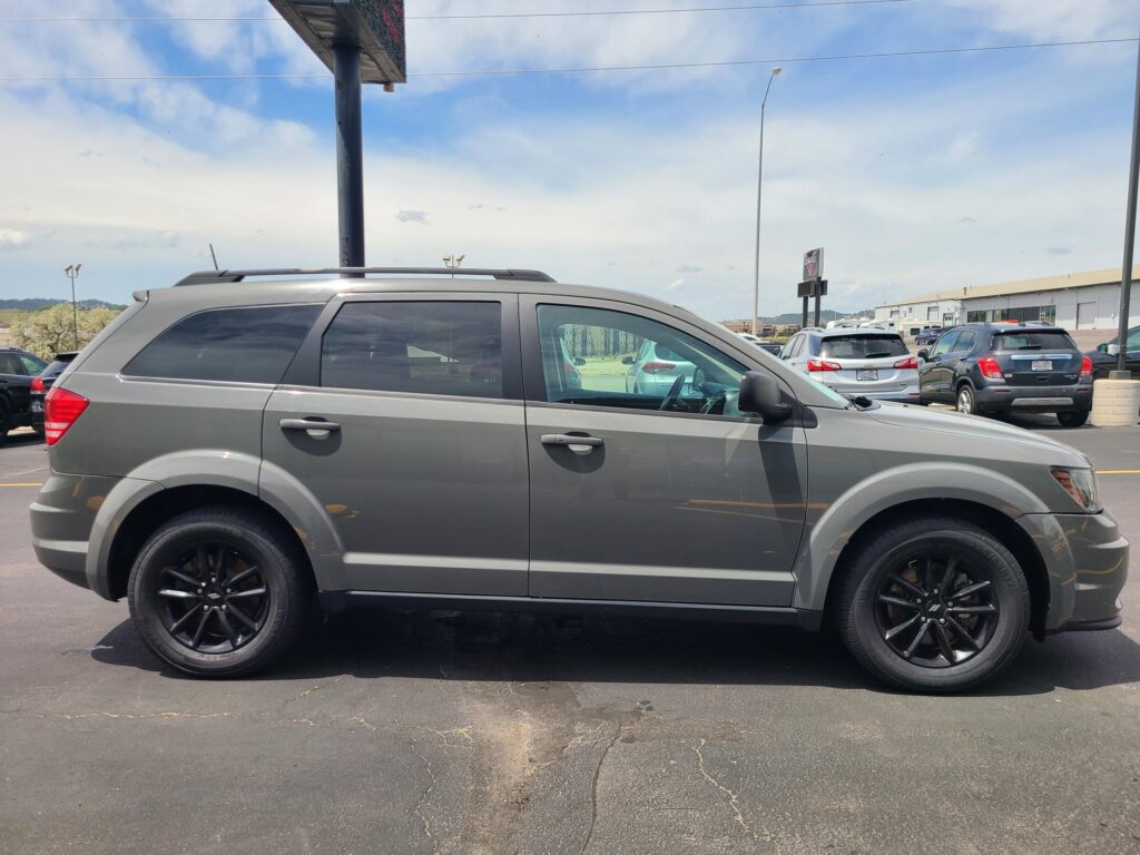 Side view of a gray SUV with black wheels parked in a lot, with other cars and a dealership-style building in the background under a blue sky.