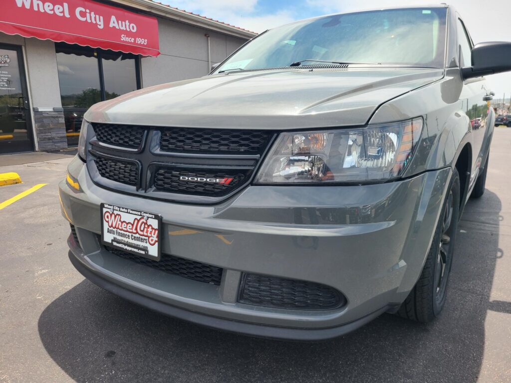 Gray Dodge van parked in front of Wheel City Auto dealership with red sign reading 'Wheel City Auto' and 'Since 1993' visible. Front grille and headlight close-up.