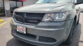 Gray Dodge van parked in front of Wheel City Auto dealership with red sign reading 'Wheel City Auto' and 'Since 1993' visible. Front grille and headlight close-up.