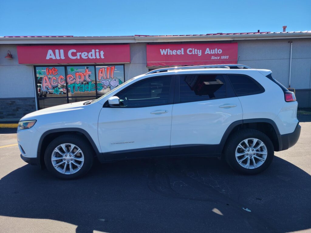 White SUV parked in front of a storefront with pink signs reading 'All Credit' and 'Wheel City Auto Since 1993'; front window mural says 'We Accept All Trades'.