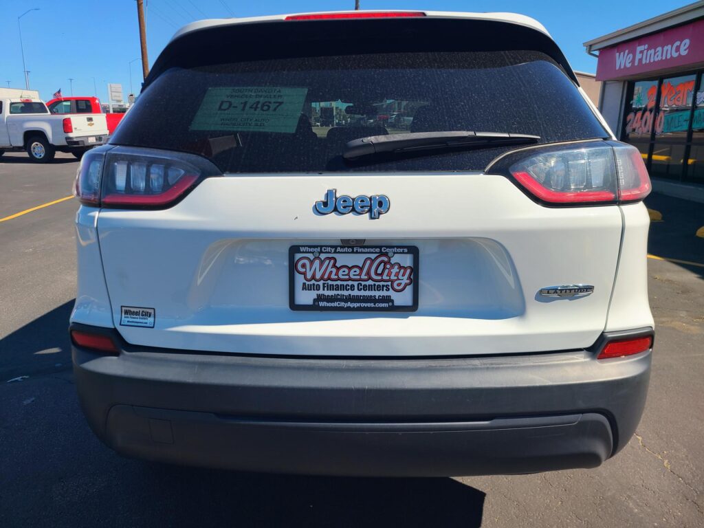 Rear view of a white Jeep Latitude at a car dealership lot with a window sticker visible in the rear window and a 'We Finance' sign on the building