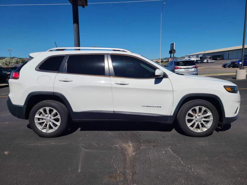 Side view of a white Jeep Cherokee SUV with roof rails parked in a lot, visible windows and wheels