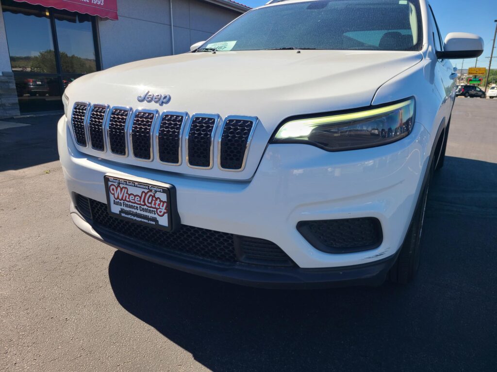 Front-right three-quarter view of a white Jeep SUV in a dealership lot, showing the seven-slot grille and illuminated headlight with a Wheel City license plate frame.