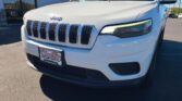 Front-right three-quarter view of a white Jeep SUV in a dealership lot, showing the seven-slot grille and illuminated headlight with a Wheel City license plate frame.