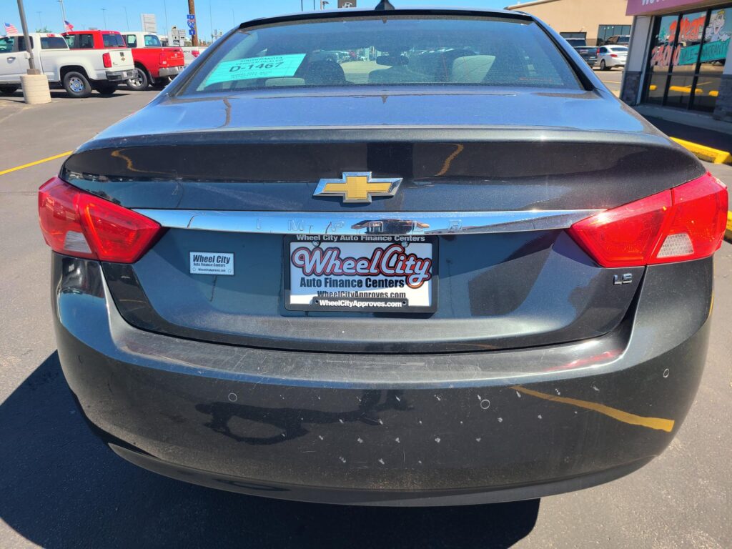 Rear view of a blue Chevrolet sedan in a dealership lot, with Wheel City Auto Finance Centers plate frame and teal window sticker visible on the rear window.