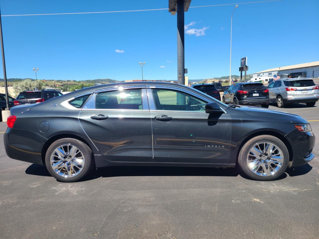 Side view of a gray Chevrolet Impala in a car dealership lot with other vehicles and a clear blue sky