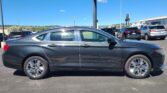 Side view of a gray Chevrolet Impala in a car dealership lot with other vehicles and a clear blue sky