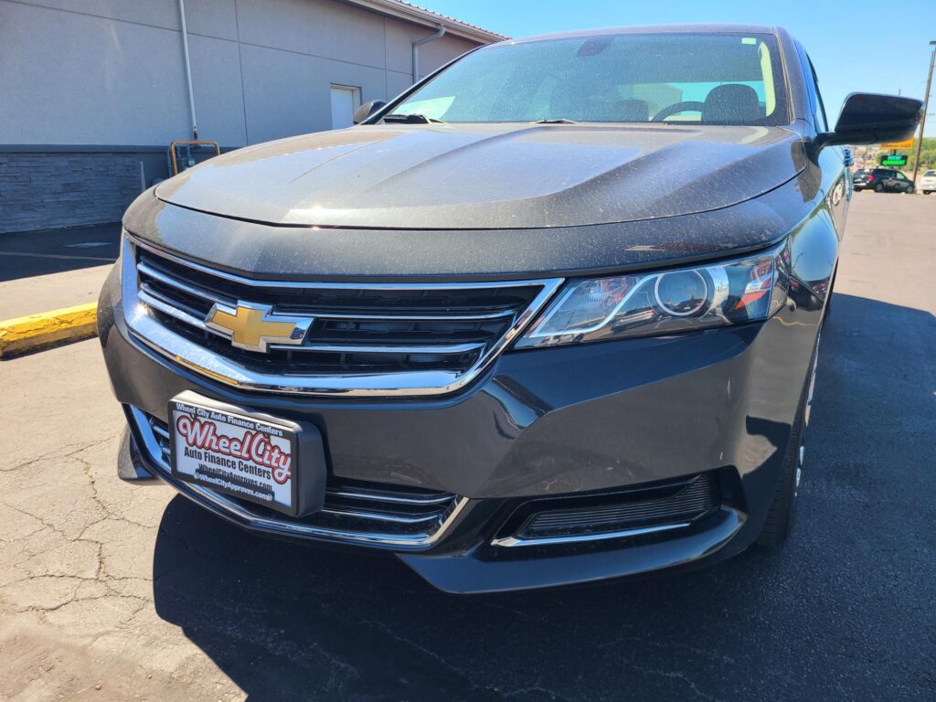 Front of a dark Chevy SUV with chrome grille, partial windshield, and a dealer plate reading 'Wheel City Auto Finance Centers' visible on the bumper.