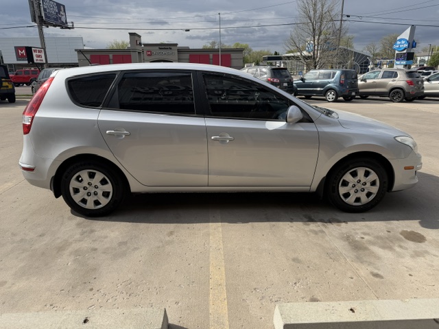 Side view of a silver minivan parked in a parking lot, with other cars and a strip mall in the background.