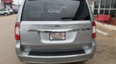Rear view of a silver Chrysler minivan at a car dealership, with a Wheel City license plate frame and a red bench outside the building.