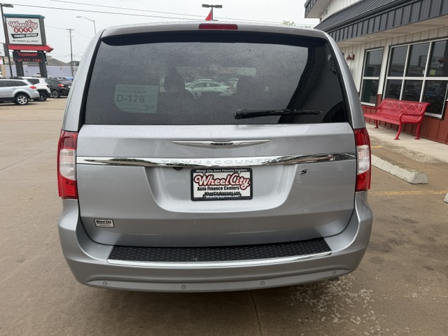 Rear view of a silver Chrysler minivan at a car dealership, with a Wheel City license plate frame and a red bench outside the building.