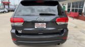 Rear view of a dark gray Jeep Grand Cherokee Laredo 4x4 at a dealership, with a Wheel City license plate frame and storefronts in the background.