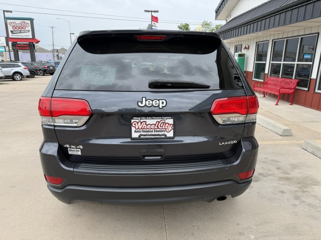 Rear view of a dark gray Jeep Grand Cherokee Laredo 4x4 at a dealership, with a Wheel City license plate frame and storefronts in the background.